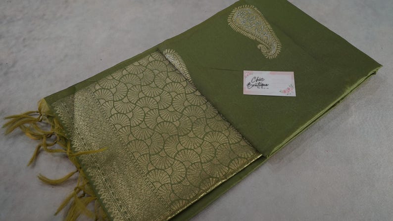 Puede incluir: Tela verde oliva con un dise&ntilde;o de cachemira dorada y un borde dorado estampado. La tela tiene una etiqueta que dice "Chic Boutique". La tela est&aacute; doblada y tiene flecos.