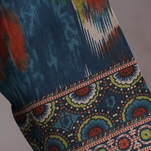 May include: Close-up of a fabric with a blue, orange, and white ikat pattern. The border features a repeating floral design in blue, green, and red on a dark blue background. The fabric appears to be a textile.