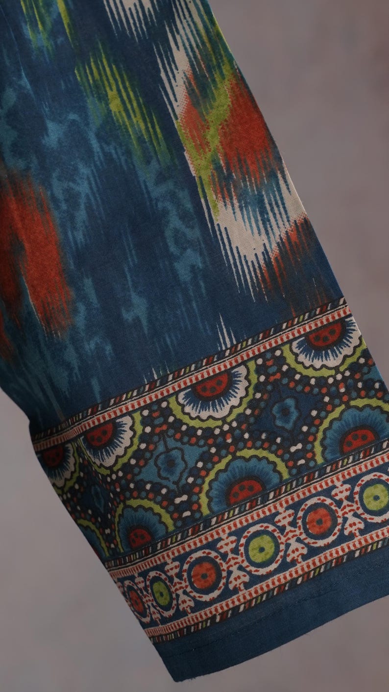 May include: Close-up of a fabric with a blue, orange, and white ikat pattern. The border features a repeating floral design in blue, green, and red on a dark blue background. The fabric appears to be a textile.
