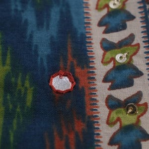 May include: Close-up of a patterned fabric with a mix of colors. The fabric features a blue, green, and orange ikat design. A silver mirror is sewn onto the fabric. The right side has a repeating pattern of blue and green figures with sequins.