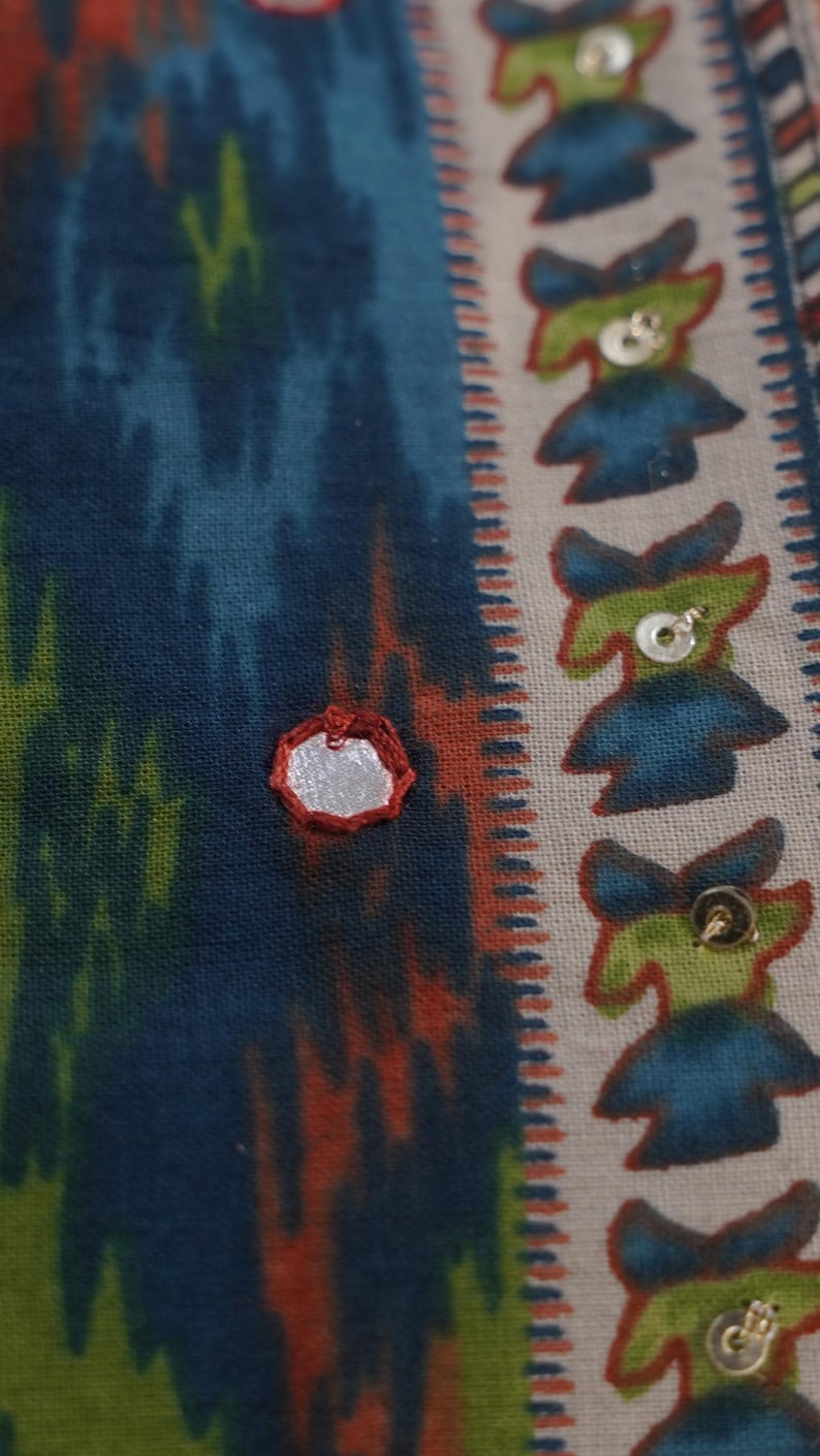 May include: Close-up of a patterned fabric with a mix of colors. The fabric features a blue, green, and orange ikat design. A silver mirror is sewn onto the fabric. The right side has a repeating pattern of blue and green figures with sequins.