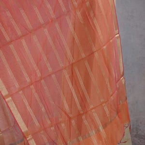 May include: A peach-coloured scarf with vertical gold stripes. The scarf has a gold border and a red trim. The fabric appears lightweight and flowing, suggesting it is made of a delicate material like silk or chiffon. The scarf is draped over a mannequin.