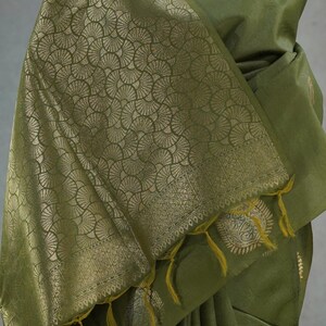 Puede incluir: Sari verde oliva con patrones dorados de abanicos y cachemir. El sari presenta un borde decorativo y borlas amarillas. La tela est&aacute; drapeada, resaltando el dise&ntilde;o y la textura del material.