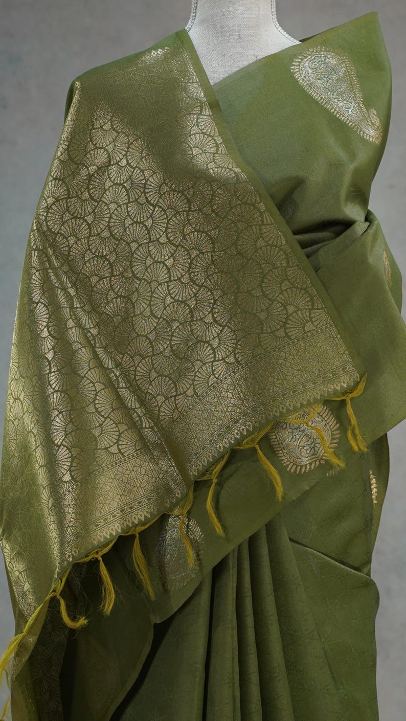 Puede incluir: Sari verde oliva con patrones dorados de abanicos y cachemir. El sari presenta un borde decorativo y borlas amarillas. La tela est&aacute; drapeada, resaltando el dise&ntilde;o y la textura del material.
