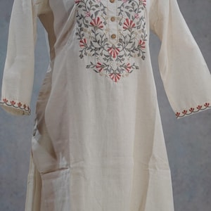 Puede incluir: Vestido t&uacute;nica crema de manga larga con bordado floral en rojo, gris y verde alrededor del cuello y los pu&ntilde;os. El vestido tiene cuello redondo y botones de madera en la parte delantera.