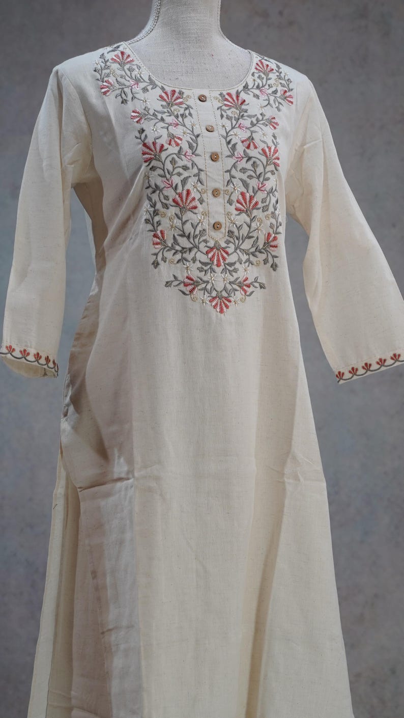 Puede incluir: Vestido t&uacute;nica crema de manga larga con bordado floral en rojo, gris y verde alrededor del cuello y los pu&ntilde;os. El vestido tiene cuello redondo y botones de madera en la parte delantera.