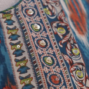 May include: Close-up of a patterned fabric with a blue, white, red, and green design. The fabric features a repeating pattern of small figures and circular motifs, embellished with small, round, reflective sequins. The overall style is reminiscent of ethnic textiles.