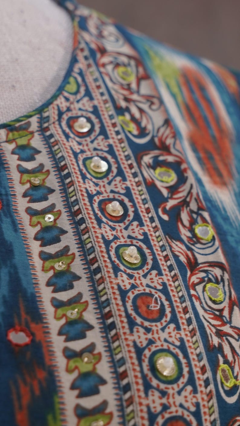 May include: Close-up of a patterned fabric with a blue, white, red, and green design. The fabric features a repeating pattern of small figures and circular motifs, embellished with small, round, reflective sequins. The overall style is reminiscent of ethnic textiles.