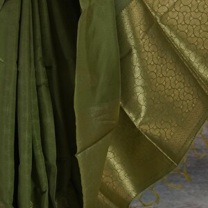 Puede incluir: Sari de seda verde oliva con un borde dorado detallado. El sari presenta un patr&oacute;n sutil en toda la tela y un borde dorado decorativo con un dise&ntilde;o en forma de abanico. La tela se drapea elegantemente, mostrando su textura y brillo.