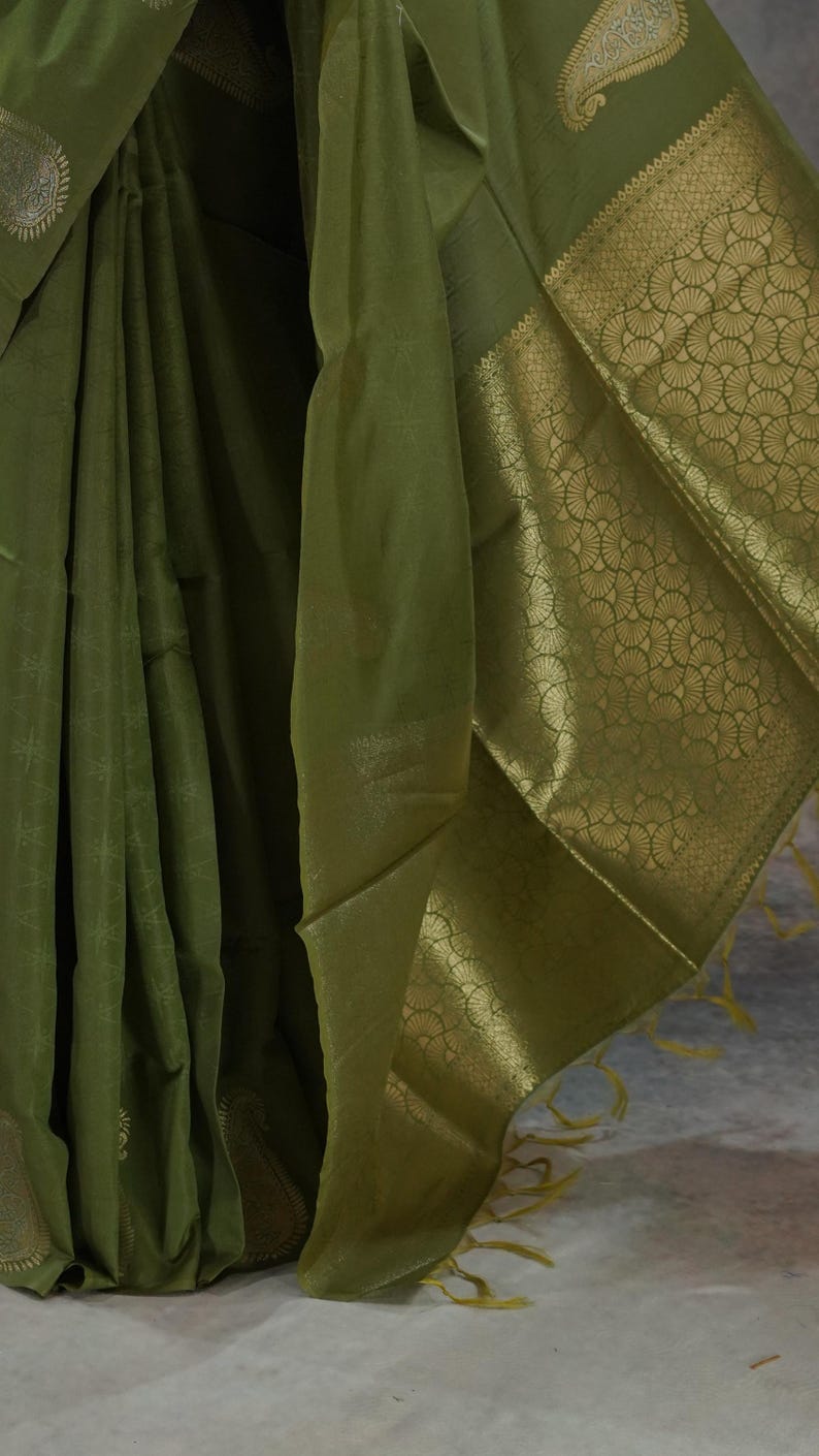 Puede incluir: Sari de seda verde oliva con un borde dorado detallado. El sari presenta un patr&oacute;n sutil en toda la tela y un borde dorado decorativo con un dise&ntilde;o en forma de abanico. La tela se drapea elegantemente, mostrando su textura y brillo.