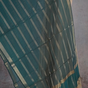 May include: A teal scarf with vertical gold stripes and gold trim. The sheer fabric drapes over a beige garment. The scarf has a rectangular shape and is likely made of a lightweight material, suitable for warmer weather.