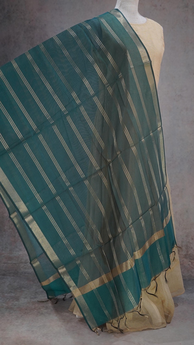 May include: A teal scarf with vertical gold stripes and gold trim. The sheer fabric drapes over a beige garment. The scarf has a rectangular shape and is likely made of a lightweight material, suitable for warmer weather.