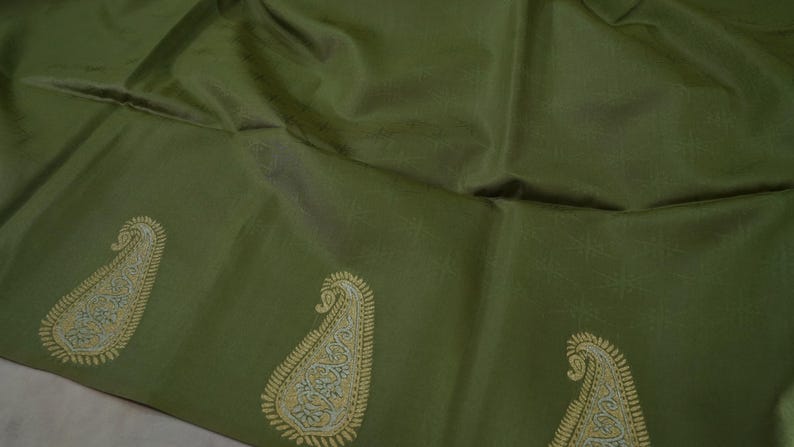 Puede incluir: Tela verde oliva con un brillo sutil. El borde inferior presenta un patr&oacute;n repetido de dise&ntilde;os de cachemira bordados en oro y azul claro. La tela tiene una textura suave y parece ser un material de alta calidad.