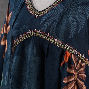 May include: A dark teal dress with a V-neckline and floral pattern. The neckline and yoke are embellished with colorful beads. The dress has a loose fit and long sleeves, with a botanical print in shades of orange and teal.