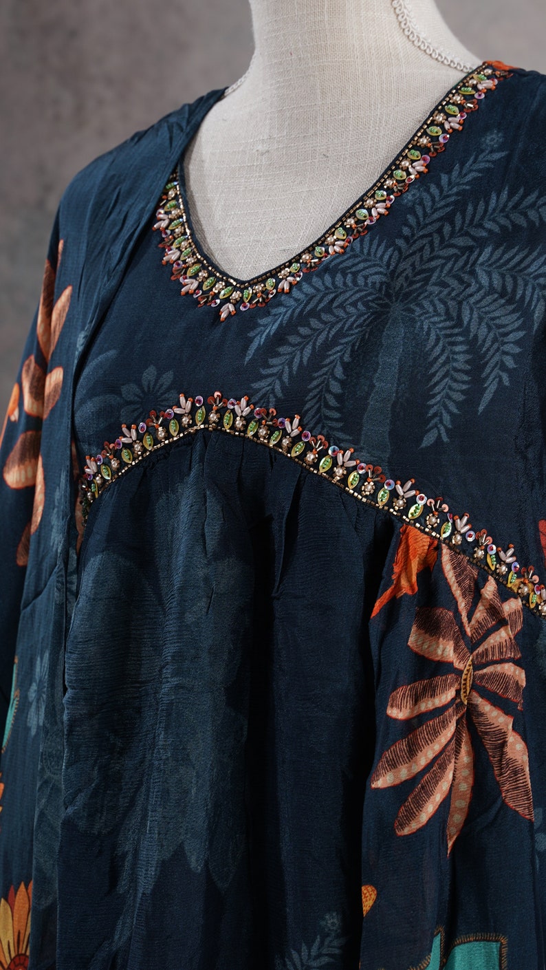 May include: A dark teal dress with a V-neckline and floral pattern. The neckline and yoke are embellished with colorful beads. The dress has a loose fit and long sleeves, with a botanical print in shades of orange and teal.