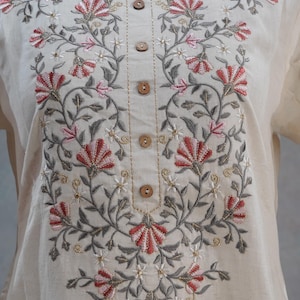 Puede incluir: T&uacute;nica color crema con intrincado bordado floral en hilo rojo, rosa, gris y dorado. El bordado forma un dise&ntilde;o sim&eacute;trico alrededor del cuello y por el frente, con botones de madera. La t&uacute;nica se exhibe en un maniqu&iacute;.