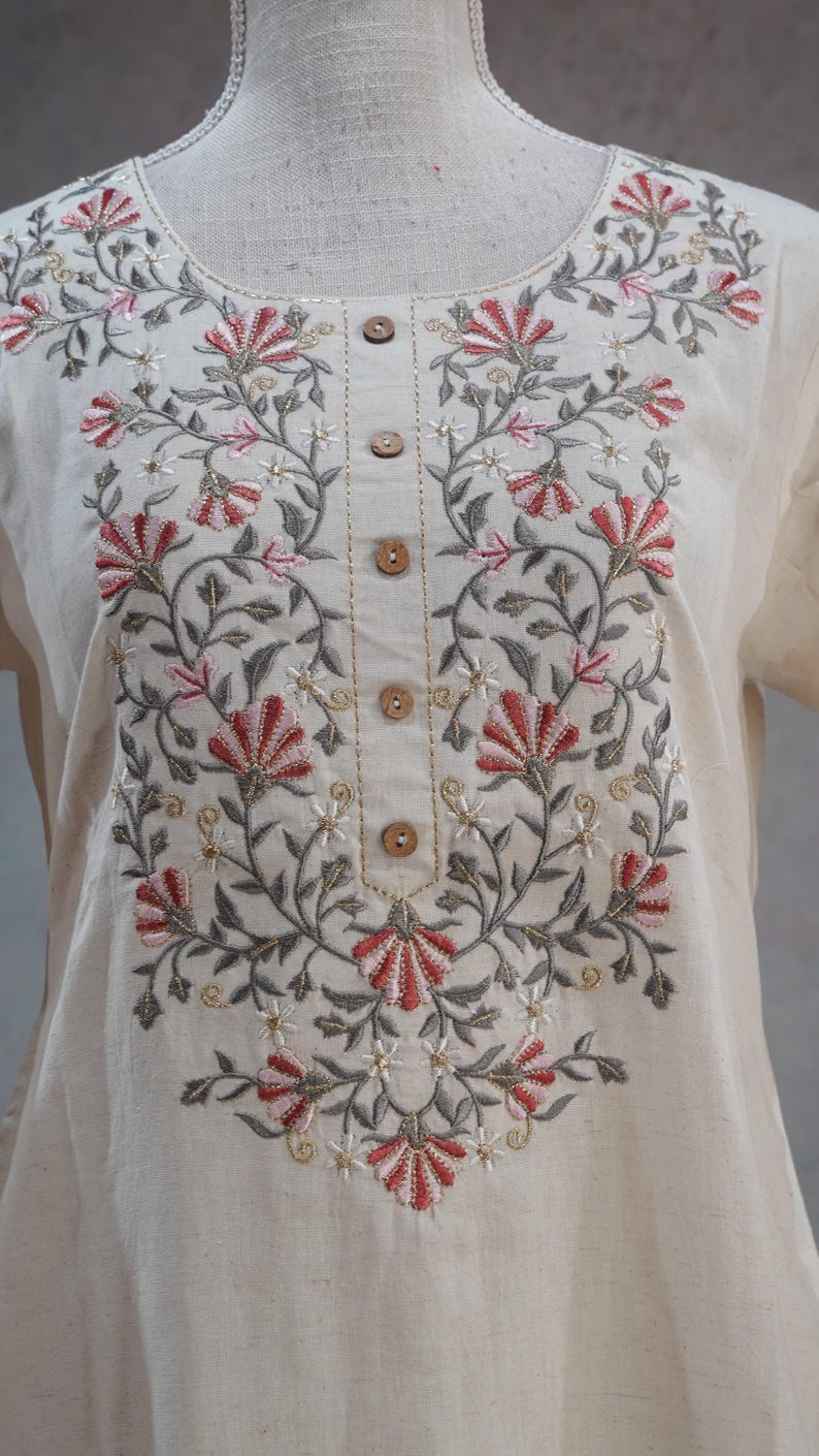 Puede incluir: T&uacute;nica color crema con intrincado bordado floral en hilo rojo, rosa, gris y dorado. El bordado forma un dise&ntilde;o sim&eacute;trico alrededor del cuello y por el frente, con botones de madera. La t&uacute;nica se exhibe en un maniqu&iacute;.