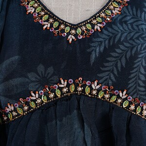 May include: Close-up of a teal-colored garment with a V-neckline and floral pattern. The neckline and waist are embellished with colorful beads and sequins. The fabric has a subtle sheen.