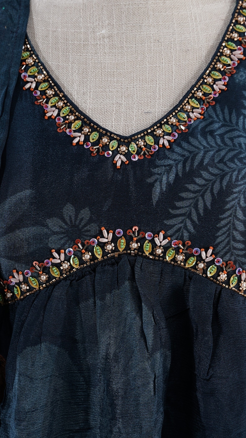 May include: Close-up of a teal-colored garment with a V-neckline and floral pattern. The neckline and waist are embellished with colorful beads and sequins. The fabric has a subtle sheen.