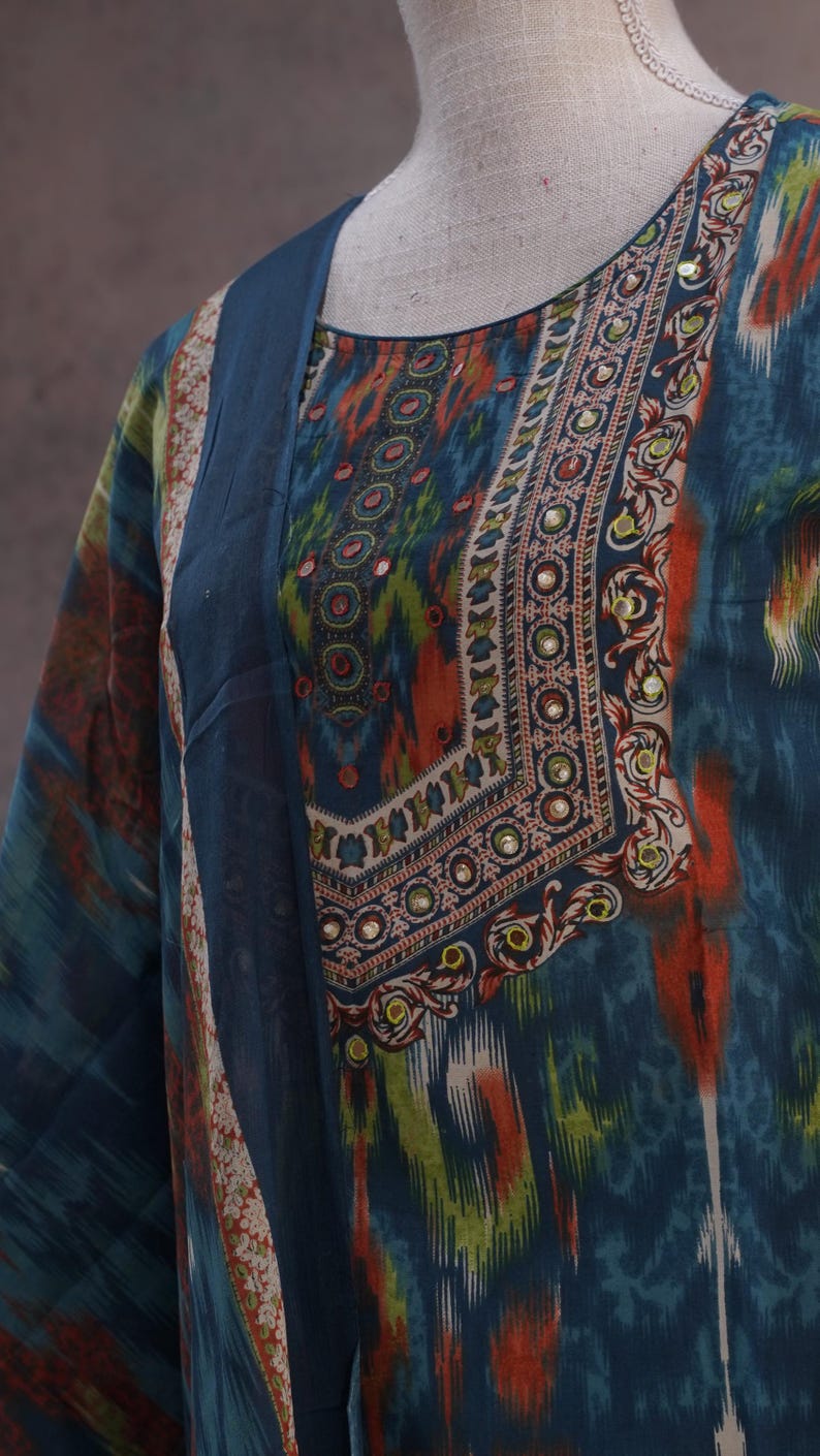 May include: A teal-colored tunic with a patterned design in orange, green, and white. The neckline and sleeves are adorned with intricate embellishments. The fabric appears to be lightweight and flowing, suitable for warm weather.