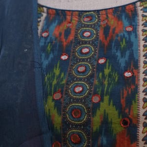 May include: Close-up of a colorful patterned fabric with a blue overlay. The fabric features a design with blue, orange, and green hues, accented with small, reflective embellishments. The garment appears to be a top or dress.