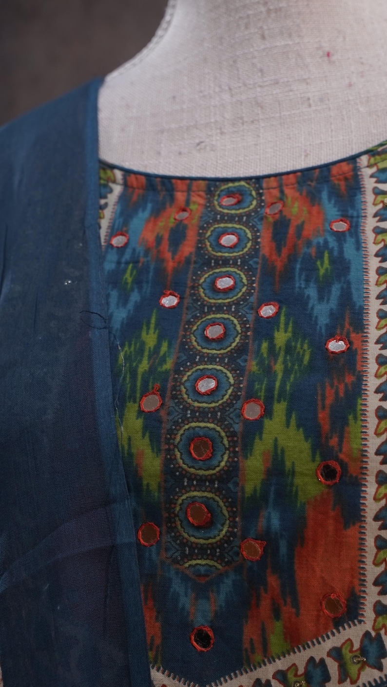 May include: Close-up of a colorful patterned fabric with a blue overlay. The fabric features a design with blue, orange, and green hues, accented with small, reflective embellishments. The garment appears to be a top or dress.