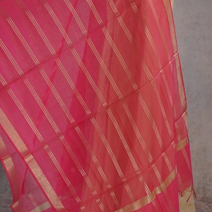 May include: A vibrant pink scarf with gold stripes and edging. The scarf is displayed, highlighting its delicate fabric and pattern. A fashion accessory.