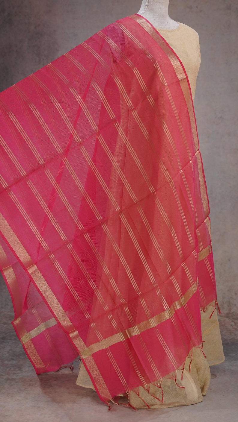 May include: A vibrant pink scarf with gold stripes and edging. The scarf is displayed, highlighting its delicate fabric and pattern. A fashion accessory.