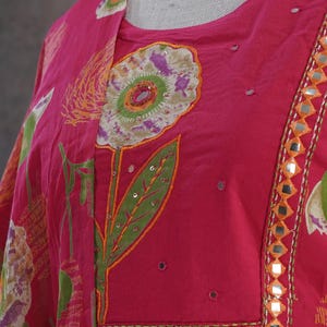 Puede incluir: Una t&uacute;nica fucsia vibrante con bordado floral. La t&uacute;nica presenta un gran dise&ntilde;o floral con hojas verdes y detalles en naranja. El escote y los bordes est&aacute;n adornados con peque&ntilde;os espejos. La tela tiene una apariencia suave y fluida.