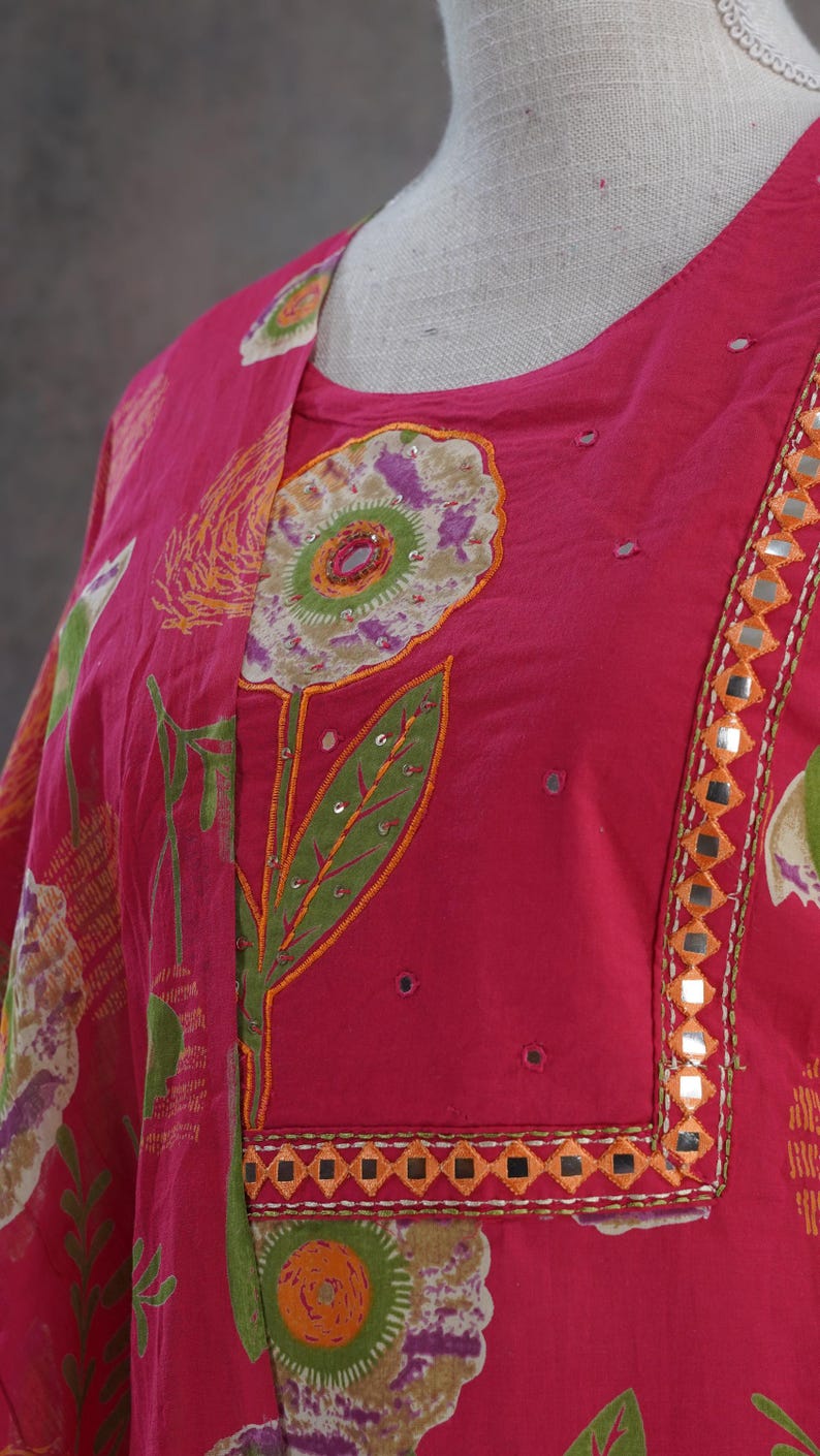Puede incluir: Una t&uacute;nica fucsia vibrante con bordado floral. La t&uacute;nica presenta un gran dise&ntilde;o floral con hojas verdes y detalles en naranja. El escote y los bordes est&aacute;n adornados con peque&ntilde;os espejos. La tela tiene una apariencia suave y fluida.