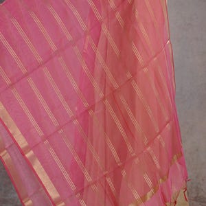 May include: A sheer pink scarf with gold vertical stripes and gold trim. The fabric is draped, revealing a glimpse of a beige garment underneath. The scarf has a delicate, airy appearance, suitable for formal or festive occasions.