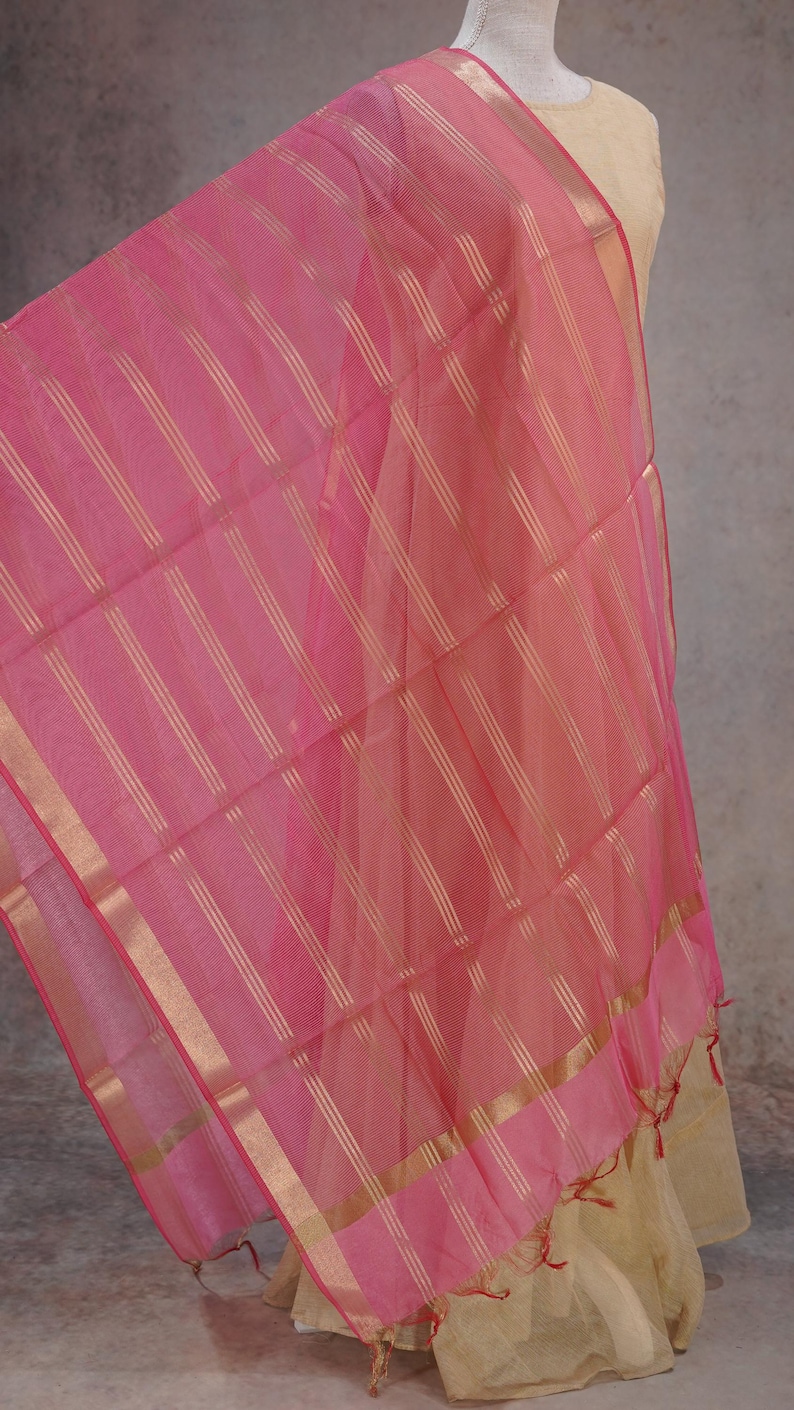 May include: A sheer pink scarf with gold vertical stripes and gold trim. The fabric is draped, revealing a glimpse of a beige garment underneath. The scarf has a delicate, airy appearance, suitable for formal or festive occasions.