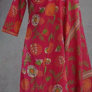 Puede incluir: Un vestido rosa vibrante con estampado floral y una bufanda a juego. El vestido presenta un dise&ntilde;o de manga larga con cuello redondo. El estampado floral incluye elementos naranjas, verdes y blancos. La bufanda tiene un borde decorativo.