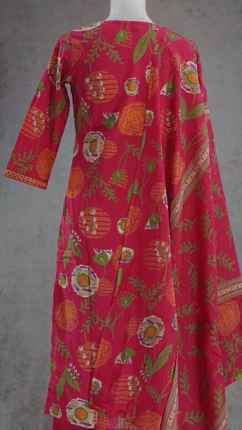 Puede incluir: Un vestido rosa vibrante con estampado floral y una bufanda a juego. El vestido presenta un dise&ntilde;o de manga larga con cuello redondo. El estampado floral incluye elementos naranjas, verdes y blancos. La bufanda tiene un borde decorativo.
