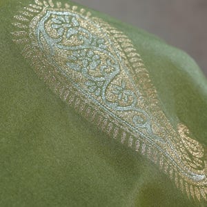 Puede incluir: Primer plano de un textil verde con un dise&ntilde;o de cachemira dorado y plateado adornado. La tela tiene una textura suave y un brillo sutil. El dise&ntilde;o se concentra a lo largo del borde de la tela.