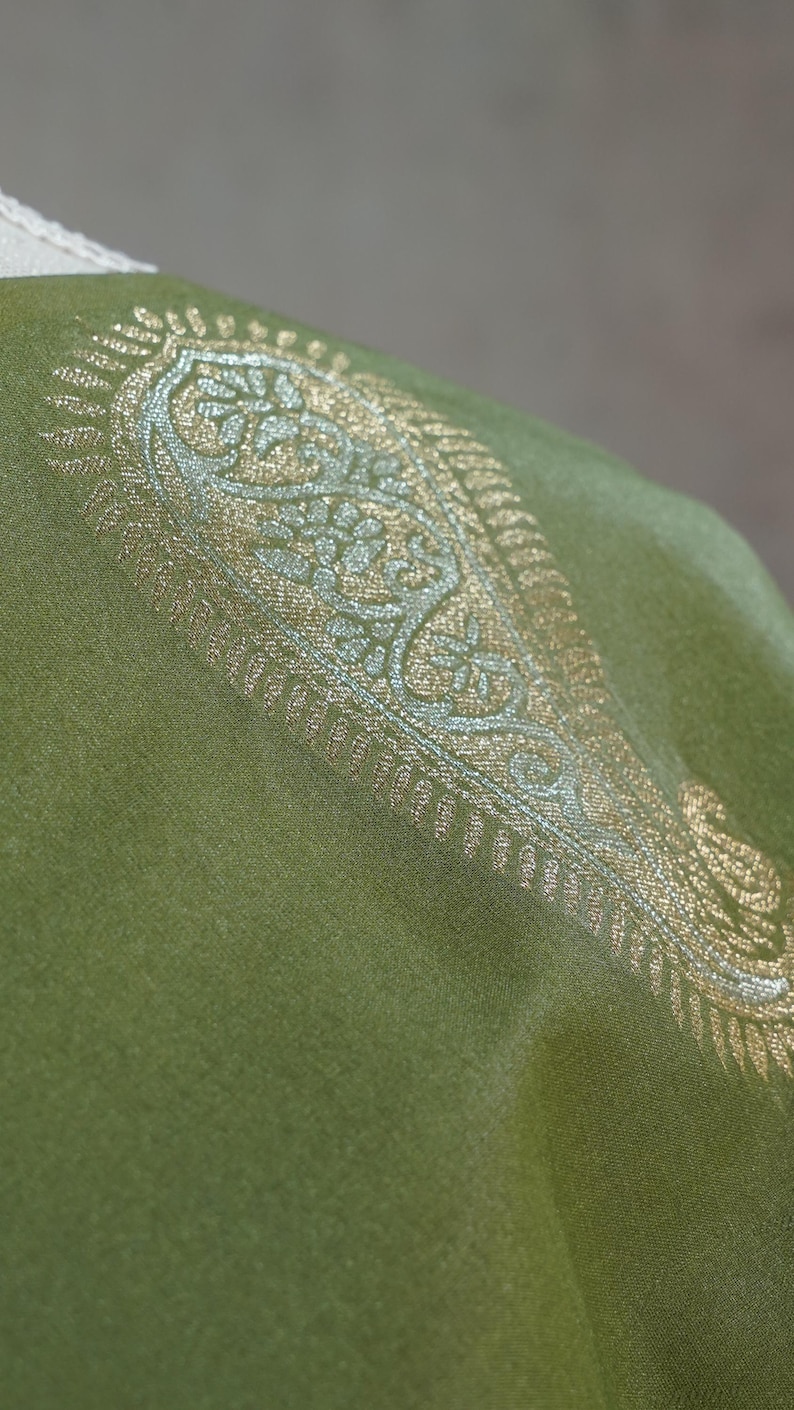 Puede incluir: Primer plano de un textil verde con un dise&ntilde;o de cachemira dorado y plateado adornado. La tela tiene una textura suave y un brillo sutil. El dise&ntilde;o se concentra a lo largo del borde de la tela.