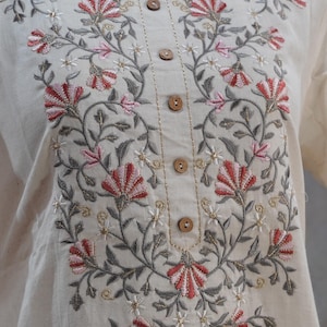 Puede incluir: T&uacute;nica color crema con intrincados bordados florales en tonos rojos, rosas, grises y dorados. El bordado adorna el escote y el frente, complementado con botones de madera. La tela parece ser lino o algod&oacute;n texturizado.