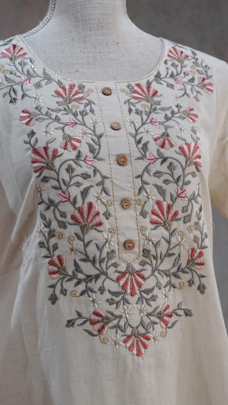 Puede incluir: T&uacute;nica color crema con intrincados bordados florales en tonos rojos, rosas, grises y dorados. El bordado adorna el escote y el frente, complementado con botones de madera. La tela parece ser lino o algod&oacute;n texturizado.