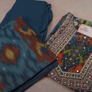 May include: A teal-colored, three-piece outfit featuring a tunic, pants, and a scarf. The tunic has intricate embroidery and mirror embellishments. The scarf and tunic have a colorful ikat pattern in shades of teal, orange, and yellow.