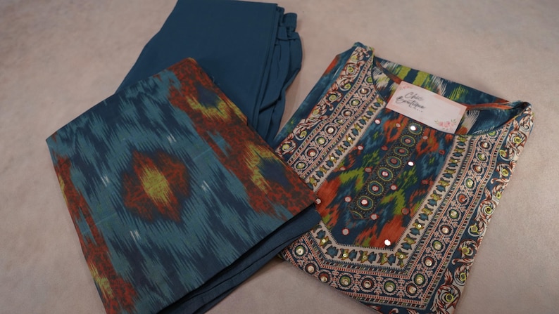 May include: A teal-colored, three-piece outfit featuring a tunic, pants, and a scarf. The tunic has intricate embroidery and mirror embellishments. The scarf and tunic have a colorful ikat pattern in shades of teal, orange, and yellow.