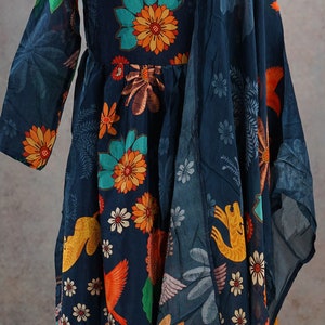 May include: A navy blue dress with a floral and bird print. The dress features long sleeves and a flowing silhouette. The print includes orange, yellow, and teal flowers and birds. A matching scarf drapes over the shoulder.