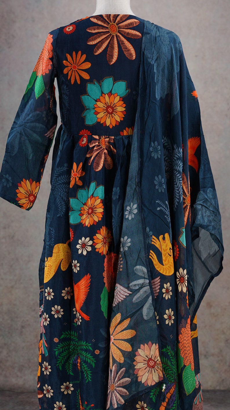 May include: A navy blue dress with a floral and bird print. The dress features long sleeves and a flowing silhouette. The print includes orange, yellow, and teal flowers and birds. A matching scarf drapes over the shoulder.