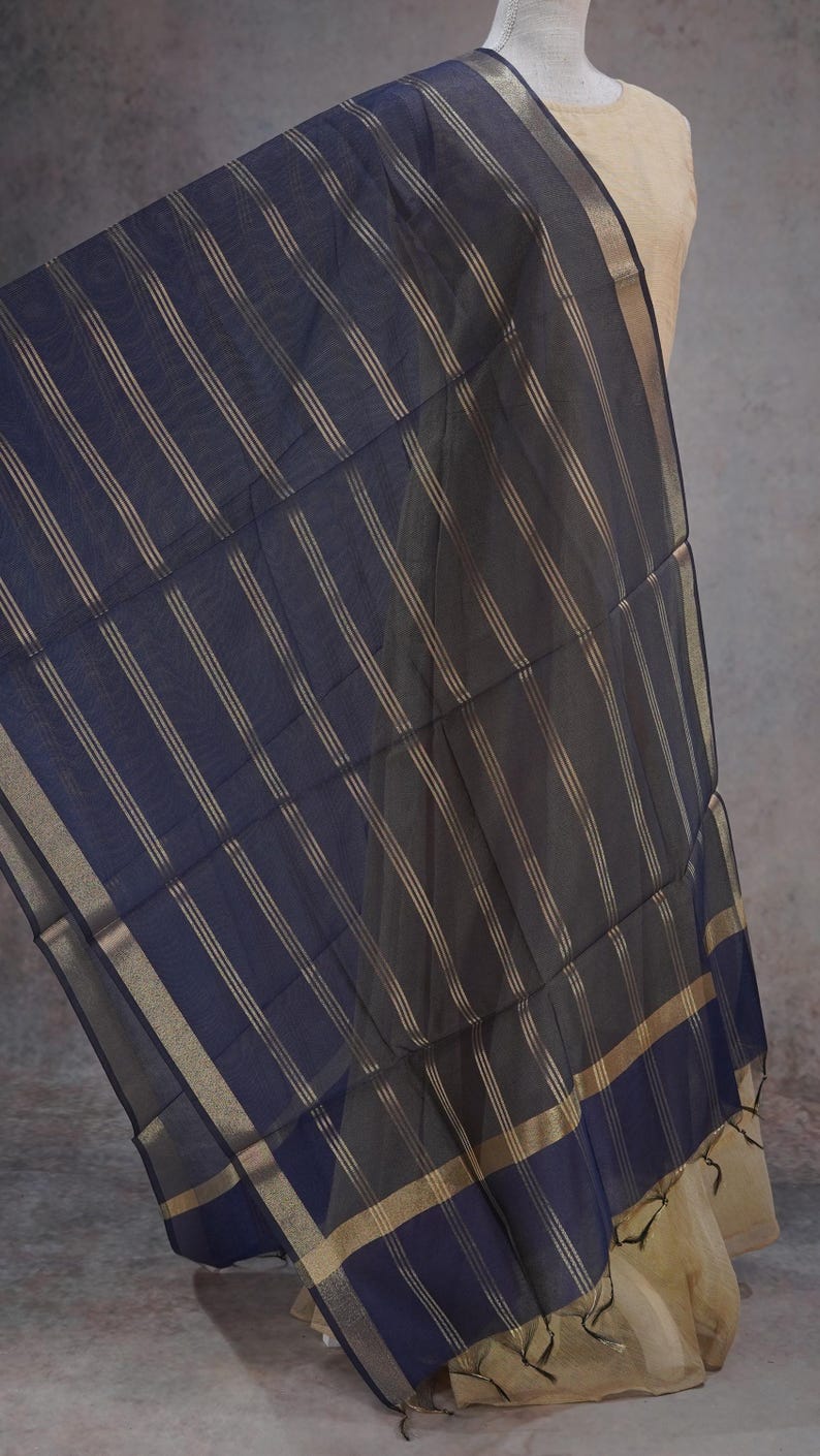 May include: A navy blue scarf with gold vertical stripes. The scarf has a gold border and is draped over a mannequin. The fabric appears lightweight and flowing, suitable for warmer weather.