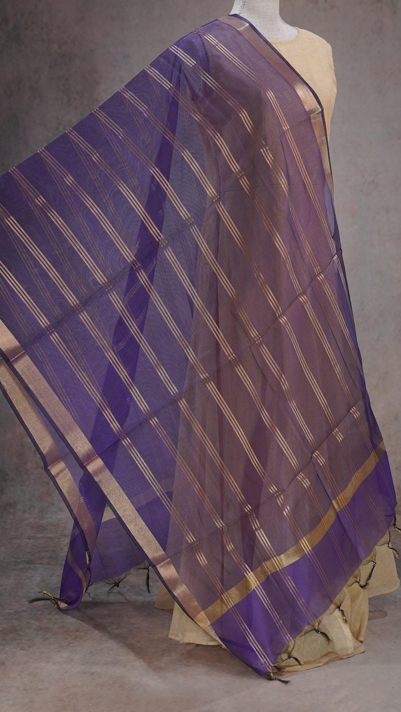 May include: A sheer purple scarf with gold stripes and trim. The scarf is draped, showcasing its flowing fabric and design. The scarf is a fashion accessory.