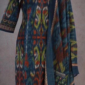 May include: A long-sleeved, patterned dress and matching scarf in shades of blue, orange, and green. The fabric features an ikat design. The dress has a straight cut, and the scarf drapes over the shoulder. The trim on the sleeves and scarf has a detailed border.