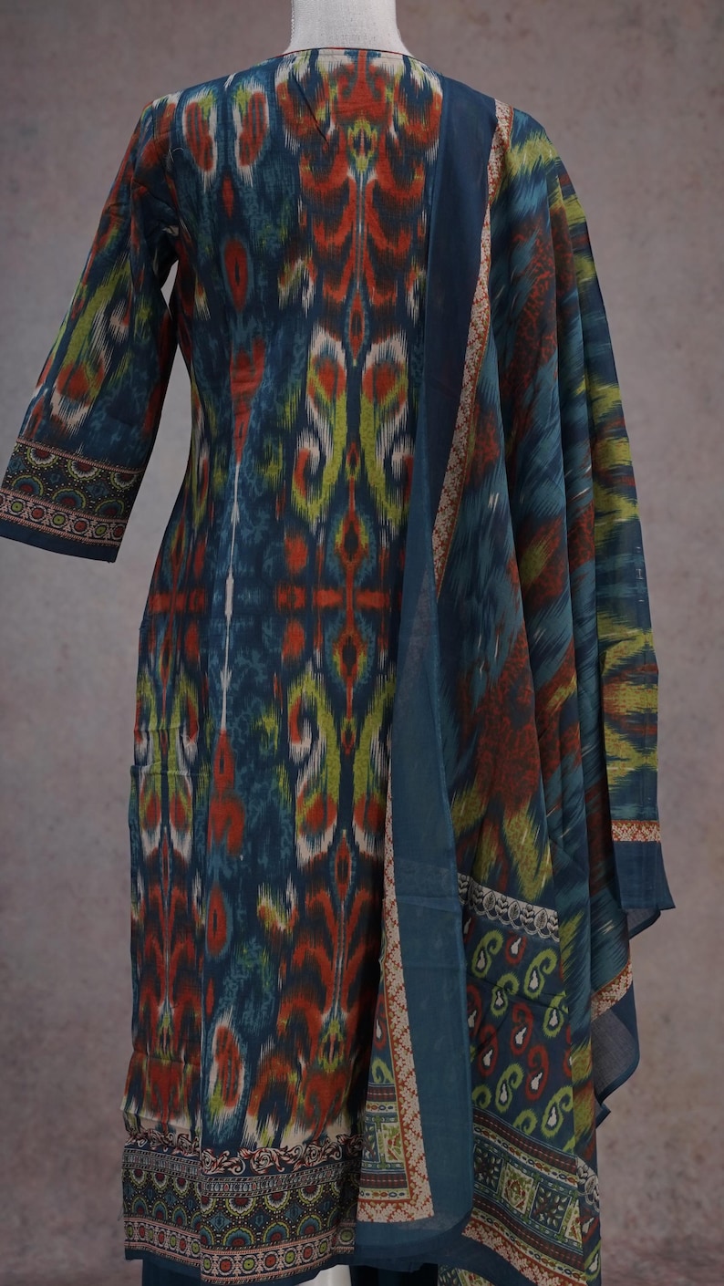 May include: A long-sleeved, patterned dress and matching scarf in shades of blue, orange, and green. The fabric features an ikat design. The dress has a straight cut, and the scarf drapes over the shoulder. The trim on the sleeves and scarf has a detailed border.
