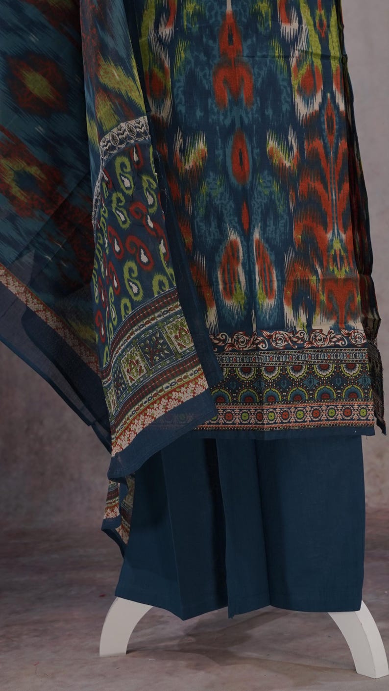 May include: A teal-colored ensemble featuring a patterned top and matching pants. The top has a vibrant ikat-style design in shades of teal, orange, and yellow. The pants are a solid teal color. The outfit includes a patterned scarf.