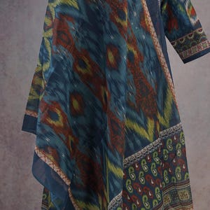 May include: A flowing, patterned garment in shades of blue, green, and red. The fabric features an ikat design, with a border of paisley and floral patterns. The sleeves are three-quarter length, and the neckline is adorned with intricate embroidery.