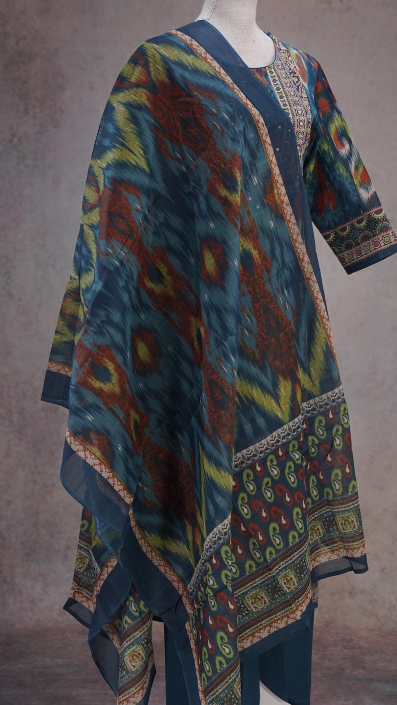 May include: A flowing, patterned garment in shades of blue, green, and red. The fabric features an ikat design, with a border of paisley and floral patterns. The sleeves are three-quarter length, and the neckline is adorned with intricate embroidery.