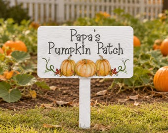 May include: A rectangular white garden sign with a wood grain texture featuring the black text 'Papa's Pumpkin Patch' above an illustration of three orange pumpkins with green vines and small colorful autumn leaves, mounted on a white stake.
